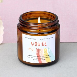 Può includere: Una candela accesa alla lavanda in un barattolo di vetro marrone. L'etichetta recita "YOU'RE my HERO" con un design arcobaleno. Il testo sull'etichetta dice anche "DAVID, YOU NEVER FAIL TO AMAZE ME... YOURSELF LOVE ERICA."