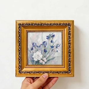 Può includere: Un dipinto a olio di iris blu e un fiore bianco in una cornice dorata quadrata e ornata. L'opera presenta pennellate spesse e materiche su uno sfondo chiaro e tenue. Il testo QURESHARTSHOP è visibile nella parte inferiore dell'immagine.