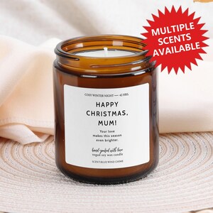 Può includere: Una candela in barattolo di vetro marrone con un'etichetta bianca che recita "BUON NATALE, MAMMA!". La candela è etichettata "COSY WINTER NIGHT - 42 HRS." e "SCENT: BLUE WIND CHIME". Un grafico rosso indica "MULTIPLE SCENTS AVAILABLE".