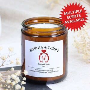 Op de afbeelding: Een bruine glazen kaarsenpot met een wit etiket met de namen "SOPHIA & TERRY" en twee pinguïn illustraties. Het etiket bevat ook de tekst "Our First Home 2024" en "HAND POURED WITH LOVE". Een rode ster geeft aan dat er meerdere geuren beschikbaar zijn.