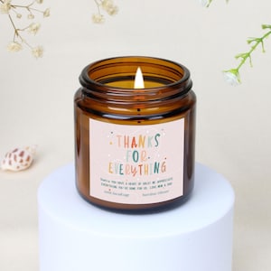 Könnte beinhalten: Eine brennende braune Glas-Kerze mit dem Text "THANKS FOR EVERYTHING" auf einem Etikett. Die Kerze steht auf einem weißen Sockel. Das Etikett enthält auch den Text "Sophia, du hast ein goldenes Herz! Wir schätzen alles, was du für uns getan hast. Love Mom & Dad".