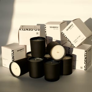 May include: A collection of black soy wax candles in matte black containers, with white labels and boxes. The boxes and labels feature the text "SO.GENTLY" and "NATURAL SOY WAX CANDLE". The candles have a white wax surface.