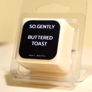 May include: A white wax melt in a clear plastic container with a black label that reads "SO. GENTLY BUTTTERED TOAST wax melts".