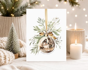 May include: A white greeting card featuring a watercolor illustration of a clear Christmas ornament hanging from a gold ribbon. Inside the ornament is a nativity scene with Mary, Joseph, and baby Jesus. Green foliage surrounds the ornament with the text Merry Christmas.