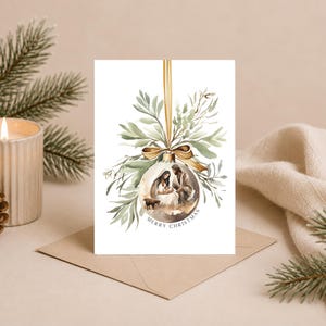 May include: A white greeting card featuring a watercolour illustration of a Christmas ornament hanging from a gold ribbon. The ornament contains a nativity scene with figures and a small animal. Green foliage surrounds the ornament, and the text Merry Christmas is printed below it.