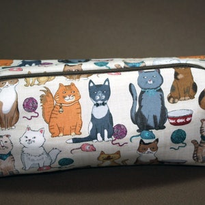 May include: A white canvas zippered pouch with a colorful print of cats. The cats are in various poses and colors, including orange, black, and brown. The pouch is perfect for storing small items like makeup, jewelry, or electronics.