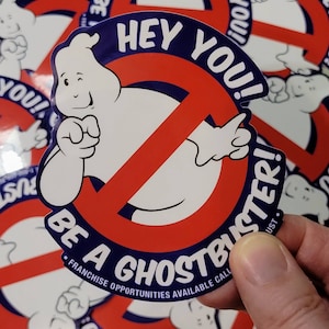 May include: A white sticker with the Ghostbusters logo, featuring a cartoon ghost within a red no-ghost symbol, and the text "HEY YOU! BE A GHOSTBUSTER!". The sticker has a blue border and the text "FRANCHISE OPPORTUNITIES AVAILABLE CALL".