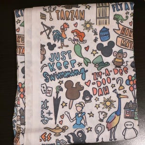 May include: White fabric with a colorful cartoon design featuring characters and phrases like "Tarzan", "Just Keep Swimming", and "Zip-A-Dee-Doo-Dah". The design includes a mermaid, Mickey Mouse, and other whimsical elements.