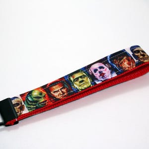May include: A fabric wrist strap key fob with a repeating pattern of classic monster movie characters. The fob has a red border and a black metal key ring. The design includes Frankenstein's monster, the Creature from the Black Lagoon, and the Mummy.