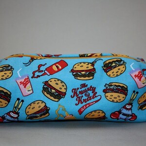 Puede incluir: Una bolsa azul con cremallera con un patrón de hamburguesas, patatas fritas y bebidas de Krusty Krab. El texto "The Krusty Krab" y "Best in Town" está impreso en la bolsa.
