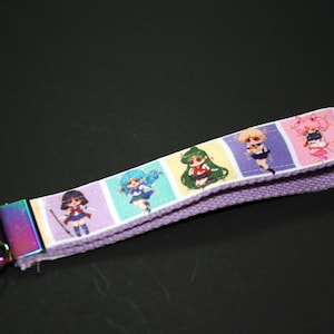 May include: A lavender wrist lanyard featuring colorful anime character illustrations. The lanyard has a metal clasp and a key ring with a rainbow finish. The design includes multiple anime characters in a repeating pattern.
