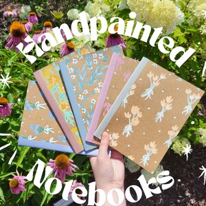 以下が含まれることがあります： 花柄の手描きノートが5冊。ノートは茶色で、白とパステルカラーの花と葉が描かれています。ノートの周りに「手描きノート」という文字が大きくスタイリッシュなフォントで書かれています。