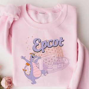 May include: A light pink sweatshirt featuring a graphic of a purple dragon with the word "Epcot" in a retro font. The design includes a stylized image of a building and stars. The sweatshirt has long sleeves and a crew neck.