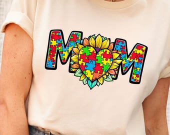 May include: A cream-colored t-shirt with the word "MOM" in a bold, colorful font. The "O" is replaced by a sunflower with a heart-shaped puzzle piece design in the center. The puzzle pieces are in various colors.
