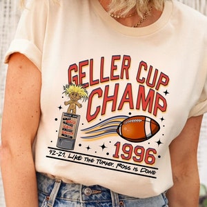May include: A cream-colored t-shirt featuring a graphic with the text GELLER CUP CHAMP 1996. The design includes a troll doll on a trophy, a football, and the text 42-21, LIKE THE TURKEY, ROSS IS DONE printed in red, blue, and black.