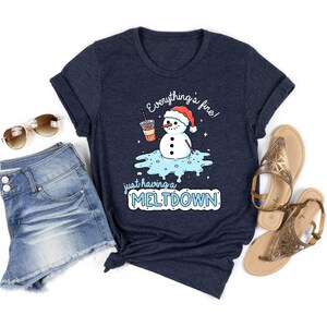 May include: Navy blue t-shirt with a snowman graphic wearing a Santa hat and holding a coffee cup. The text reads "Everything's fine! just having a MELTDOWN". The shirt is paired with denim shorts and sunglasses.