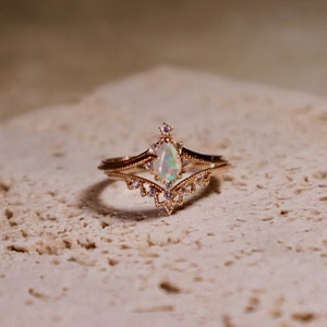 Puede incluir: Un anillo de oro con una piedra de ópalo en forma de pera rodeada de diamantes más pequeños. El anillo está colocado sobre una superficie blanca texturizada.