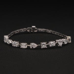 May include: A silver bracelet with a variety of diamond shapes, including emerald, pear, and square cuts. The bracelet is set against a black background, highlighting the sparkle of the gemstones. A delicate piece of jewelry.