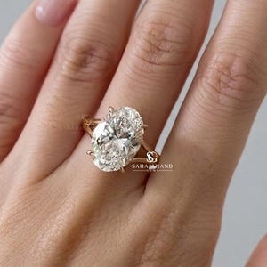 Op de afbeelding: Een gouden ring met een grote ovaal geslepen middensteen die door vier pootjes op een gespleten ringband wordt vastgehouden. Het metaal heeft een warme gele tint en de zetting bevat een klein logo met de tekst SAHAJANAND GEMS bij de basis.