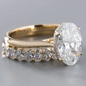 Op de afbeelding: Een gouden ringenset met een grote ovaal geslepen middensteen in een klauwzetting, voorzien van een verborgen halo van kleine diamanten. De ring is gecombineerd met een bijpassende aansluitring bezet met een rij kleine, ovaal geslepen stenen.