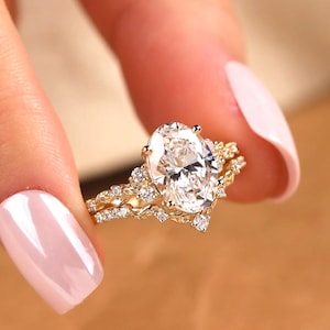 May include: A gold ring set featuring a large oval-cut centre stone surrounded by smaller round stones. The band has a detailed leaf-like texture and a V-shaped contour, with additional small stones embedded along the metal surface.