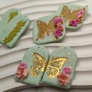 Peut inclure: Un ensemble de quatre boucles d'oreilles en céramique faites à la main avec un motif de papillon avec des accents dorés et des fleurs roses. Les boucles d'oreilles sont sur un fond vert clair.
