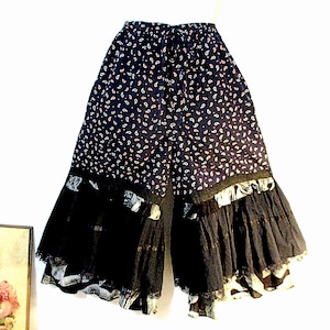 May include: Wide-leg pants with a navy blue paisley print and black lace trim. The pants have a drawstring waist and a tiered, ruffled hem. The paisley pattern includes white and pink accents.