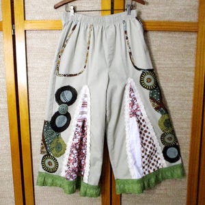 May include: Light gray culottes with decorative patches and a green fringe hem. The patches feature floral and geometric patterns in shades of brown, green, and white. The pants are hanging on a wooden hanger.