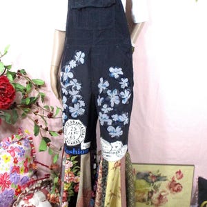 May include: Black denim overalls featuring a bib pocket, white floral embroidery on the upper legs, and circular postage stamp-style patches. The lower pant legs are flared with multi-patterned fabric panels in yellow, floral, and geometric prints.