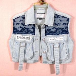 May include: A light blue denim vest featuring dark blue lace panels on the upper chest, two front pockets with hanging straps, and metal buttons. The left pocket has the text "SPÉCIALITÉ" and the right pocket has "Côté de la Rue Castiglione".