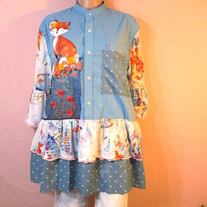May include: A patchwork tunic-style shirt with a whimsical design. The shirt features a blue gingham pattern, floral and polka dot accents, and a fox applique. The sleeves have a ruffled design, and the hemline is tiered.
