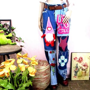 May include: Blue denim jeans featuring a brown leather belt, a pink gnome appliqué, a pink star patch, red rose embroidery, and a white lace square. The jeans include a pink rectangular patch with the text BE YOU TIFUL in white letters.