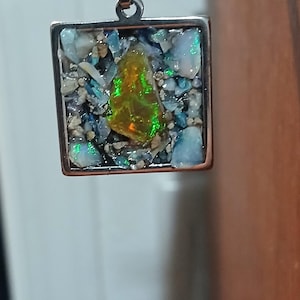 May include: Square pendant with a silver bail, featuring a mosaic of small, colorful stones and a central, iridescent, yellow-green gemstone. The pendant is set in a copper-colored frame, creating a unique jewelry piece.