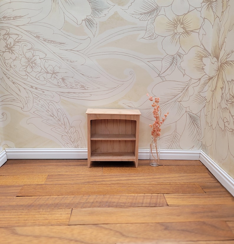 May include: A small, unfinished wooden bookshelf with two shelves. The bookshelf is set against a floral wallpaper background and sits on a wooden floor. A small glass vase with dried flowers is to the right of the bookshelf.