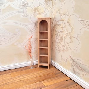 May include: A miniature wooden bookshelf with four shelves. The bookshelf is light brown and has an arched top. A small glass vase with dried flowers sits next to the bookshelf. The background features a floral wallpaper and wooden flooring.