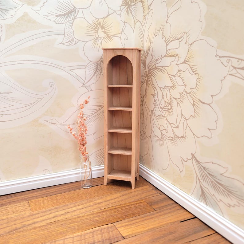 May include: A miniature wooden bookshelf with four shelves. The bookshelf is light brown and has an arched top. A small glass vase with dried flowers sits next to the bookshelf. The background features a floral wallpaper and wooden flooring.
