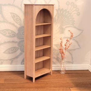 May include: A miniature wooden bookshelf with an arched top and four shelves. The natural wood finish complements the light-colored floral wallpaper in the background. A small vase with dried flowers sits nearby.