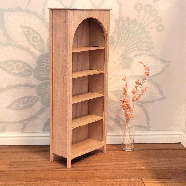 May include: A miniature wooden bookshelf with an arched top and four shelves. The natural wood finish complements the light-colored floral wallpaper in the background. A small vase with dried flowers sits nearby.