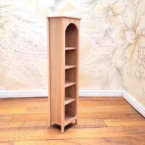 May include: A miniature wooden bookshelf with four shelves. The bookshelf has an arched top and is made of light-colored wood. It is designed to be a dollhouse or miniature display item. The bookshelf is standing on a wooden floor.