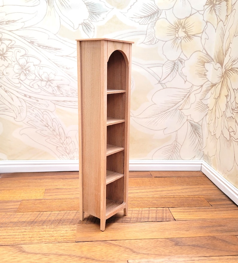 May include: A miniature wooden bookshelf with four shelves. The bookshelf has an arched top and is made of light-colored wood. It is designed to be a dollhouse or miniature display item. The bookshelf is standing on a wooden floor.