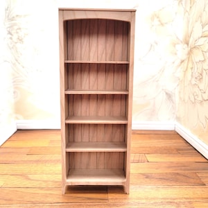 May include: A miniature wooden bookshelf with five shelves. The bookshelf is made of light-colored wood and has a simple, classic design. The top of the bookshelf has an arched shape. The shelves are evenly spaced and appear to be empty.