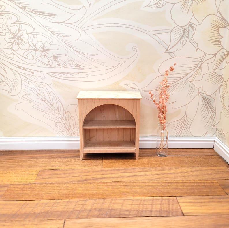 May include: A small, unfinished wooden bookshelf with two shelves and an arched opening. The miniature bookcase is made of light-colored wood and has a simple, classic design. It is a dollhouse-sized piece of furniture.