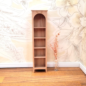 May include: A miniature wooden bookshelf with five shelves and an arched top. The bookshelf is made of light-colored wood and stands on a wooden floor. A small glass bottle with dried flowers is next to the bookshelf.