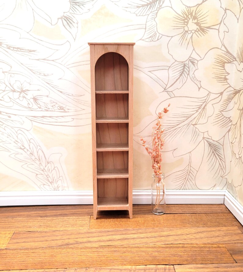 May include: A miniature wooden bookshelf with five shelves and an arched top. The bookshelf is made of light-colored wood and stands on a wooden floor. A small glass bottle with dried flowers is next to the bookshelf.