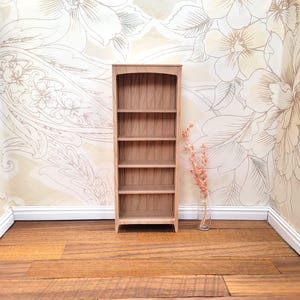 May include: A miniature wooden bookshelf with five shelves. The bookshelf is made of light-colored wood and has an arched top. The background features floral wallpaper and a vase with dried flowers. The floor is made of wooden planks.