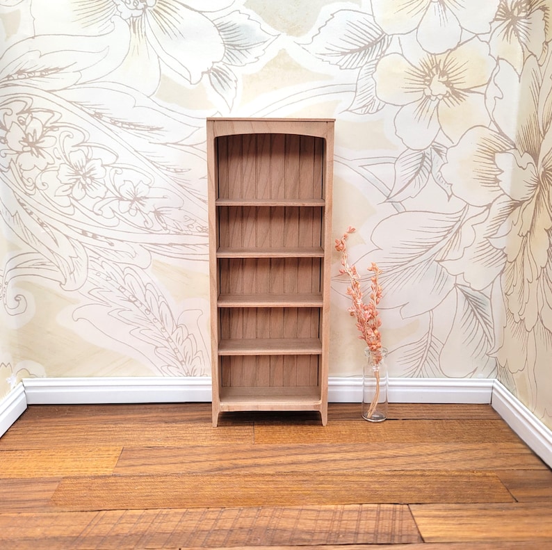 May include: A miniature wooden bookshelf with five shelves. The bookshelf is made of light-colored wood and has an arched top. The background features floral wallpaper and a vase with dried flowers. The floor is made of wooden planks.