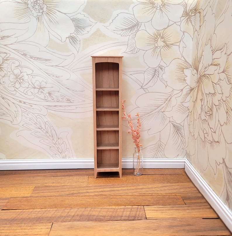 May include: A miniature wooden bookshelf with five shelves stands on a wooden floor. A small glass bottle with dried flowers is next to the bookshelf. The background features floral wallpaper in neutral tones.