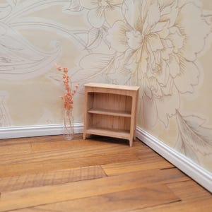 May include: A small, unfinished wooden bookshelf with two shelves. The miniature bookcase is set against a floral wallpaper background and sits on a wooden floor. A clear glass vase with dried flowers is to the left of the bookshelf.