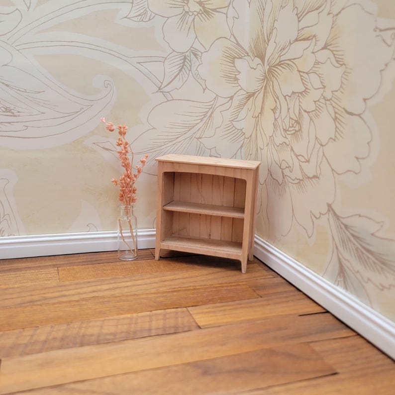 May include: A small, unfinished wooden bookshelf with two shelves. The miniature bookcase is set against a floral wallpaper background and sits on a wooden floor. A clear glass vase with dried flowers is to the left of the bookshelf.