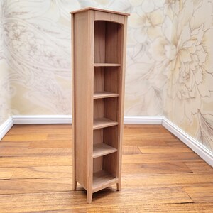 May include: A miniature wooden bookshelf with four shelves. The bookshelf is made of light-colored wood and has a tall, narrow design. The top of the bookshelf has a curved arch. The shelves are evenly spaced and appear to be empty. The bookshelf is a dollhouse or miniature furniture item.